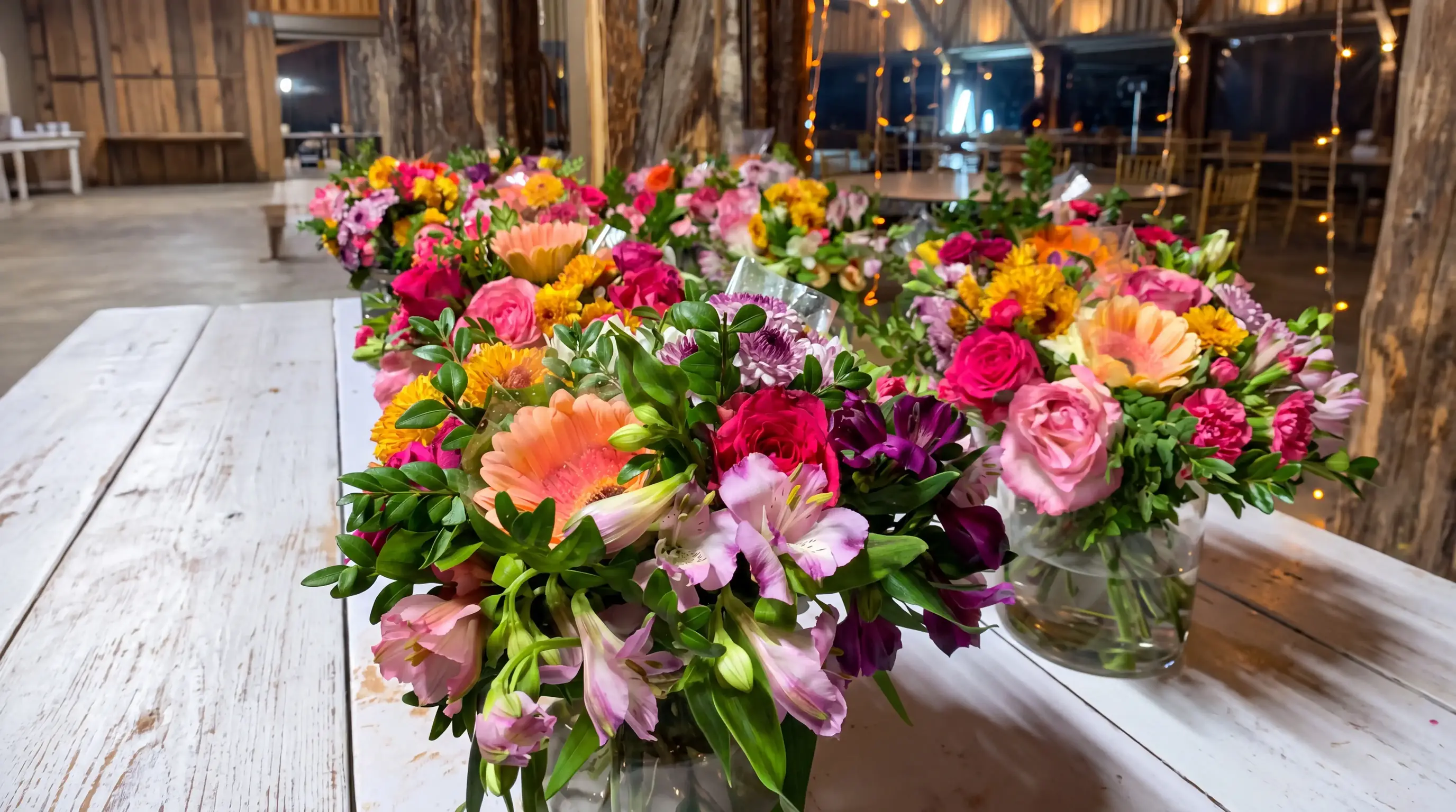Mesa de madera blanca con arreglos de gerberas, rosas, alstroemerias y claveles de colores vivos en un salón con luces cálidas
